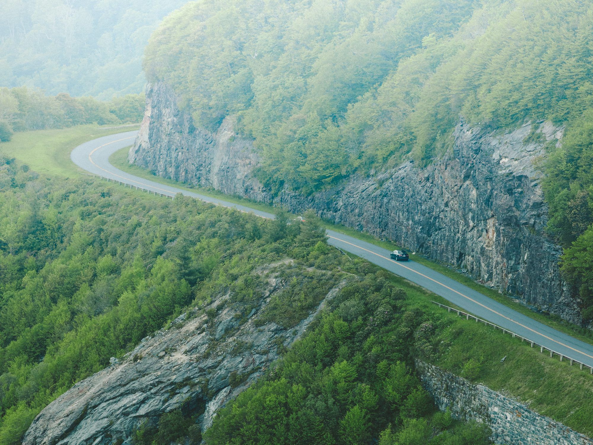 A two-lane mountain road curving along a rock cut above a forested valley, with a single car on it