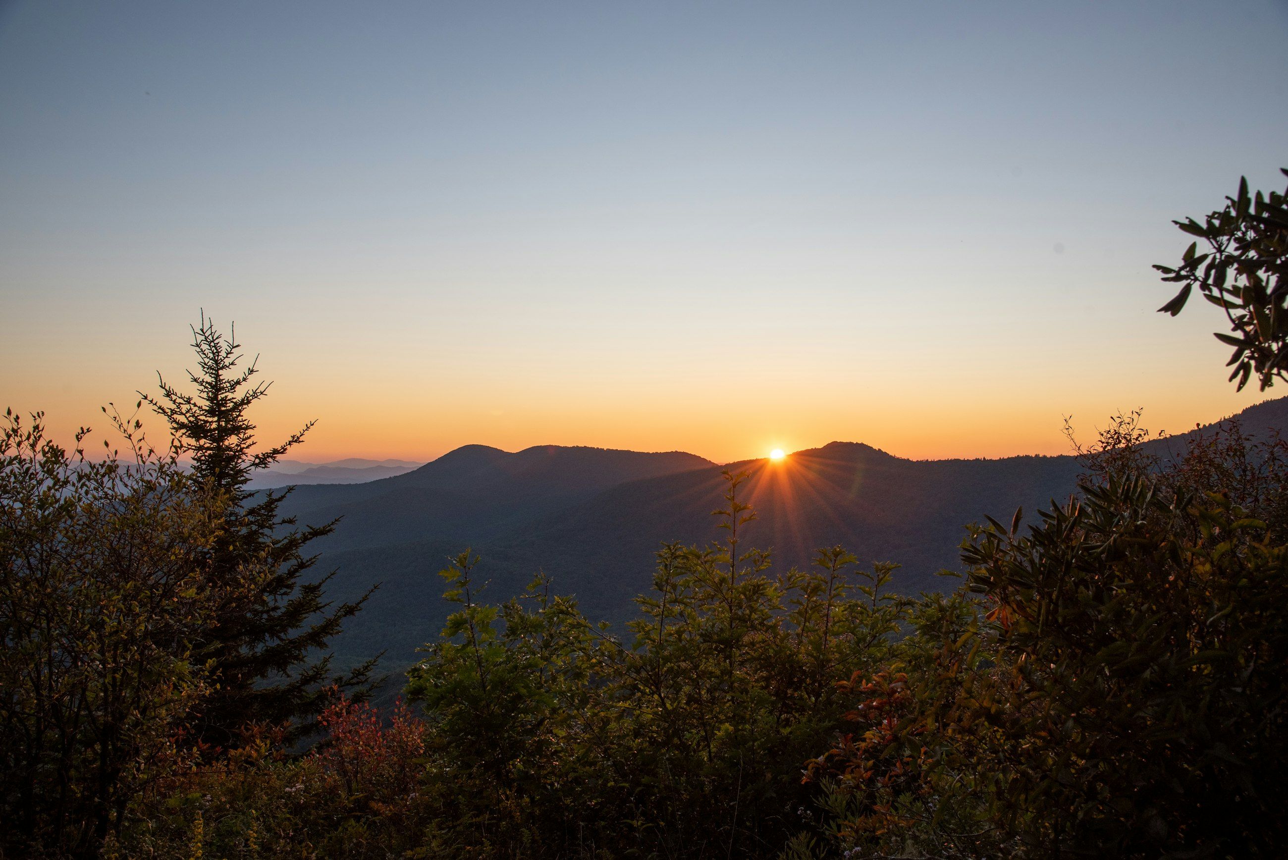 The sun setting behind a dark mountain ridgeline, with shrubs silhouetted in the foreground